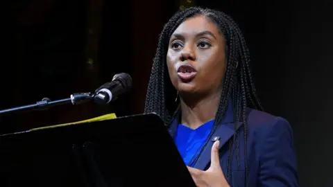 Conservative leader Kemi Badenoch, standing behind a black lectern on a stage, speaks to an audience