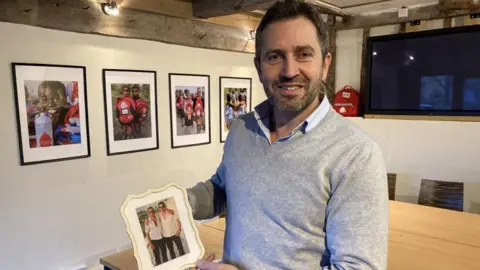 BBC Luke Simon stood in his house holding a photograph of him and his brother Piers when they were younger. There are photos of children on the wall who have been helped through his charity.