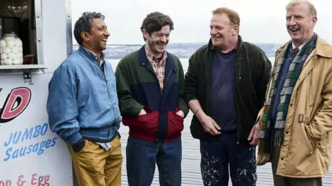 BBC / Quay Street Productions / Tom Jackson Four stars of the drama Men Up stand side-by-side and laughting next to a food van on a pier.