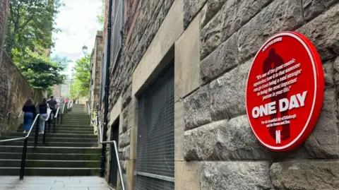 The red sign with One Day in large white letters and a quote is attached to a historic wall on the right hand side of the picture. There are stone steps rising up into the distance. There are people walking up the steps in dark clothing.