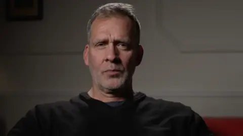 James Harvey wearing a black top in a dimly lit room, speaking to the BBC. He has short grey, white hair and stubble.