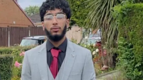 GMP Mohammad Danyaal Asghar-Ali, 19, who has black curly hair and black beard and is wearing light framed glasses, poses for a picture in a residential garden wearing a grey suit over a black collared shirt with a pink tie. 