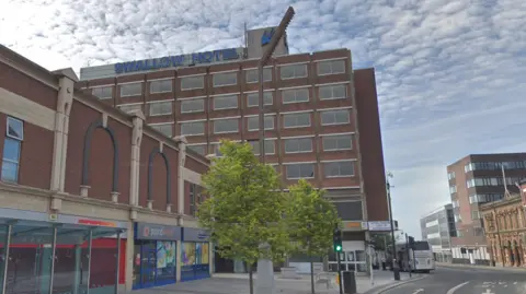 Google The Swallow Hotel is an eight-storey red-bricked hotel that stands tall over the high street shops in front of it. Blocks of uniform hotel rooms have large, square, grey windows in rows. A sign at the top is blue and reads "Swallow Hotel". To the right, the same blue glass is in the shape of a swallow bird in flight.