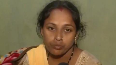 The image shows a woman with medium brown skin and dark hair pulled back. She is wearing a light-colored traditional outfit with a yellow shawl draped over one shoulder. The woman has a red bindi on her forehead and gold earrings, and she appears to be speaking or being interviewed in front of a plain, muted green background.