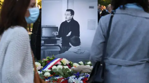 AFP Mourners pause outside the headline showing a gray and white photo of French high school teacher Samuel Paty 