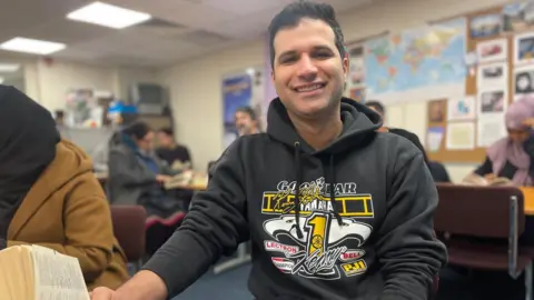 Simon Thake A smiling man in a black hoodie sits on an office chair with a map of the World behind him.