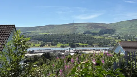 A wide shot of Re-Gen's Warrenpoint premises, taken from an elevated location in a nearby housing estate. Waste bales are stacked in piles on the harbour and a line of lorry trailers are parked nearby. The roofs of two houses are partially visible in the foreground and Carlingford Lough and green hills are in the background.