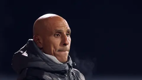 Dino Maamria watching a match with a big hooded puffa jacket zipped up to the top. His breath is visible 