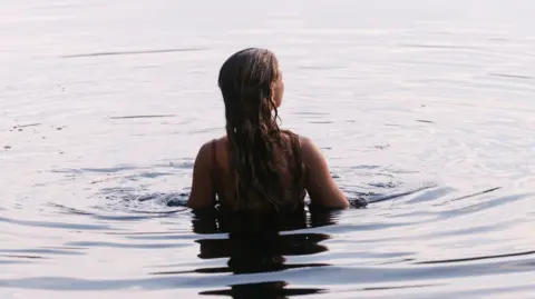 A woman in water up to her mid-back with her back to the camera. She has long, dark hair past her shoulders and the water ripples around her