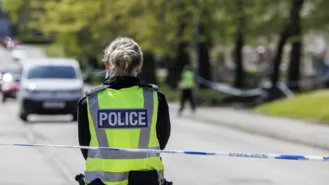 A female police officer is standing with her back to the camera, wearing a yellow hi-vis tabard saying 'POLICE'. There is police tape behind her. It is a sunny day and more police and a white van can be seen - out of focus - in the distance.