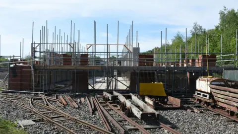 Apedale Valley Light Railway A series of railway tracks lead up to a construction site which comprises scaffolding and a brick-built structure. There are various pieces of railway equipment on and alongside the tracks.