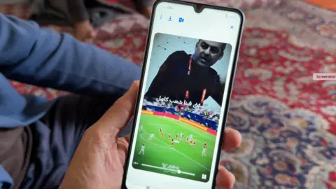 Huda holding a mobile phone where trams are playing football, with the image of the head coach displayed above the stands