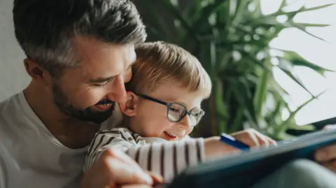 Getty Images A young boy sitting on a man's knee. They are sitting in a home and drawing on a tablet. They are both smiling.