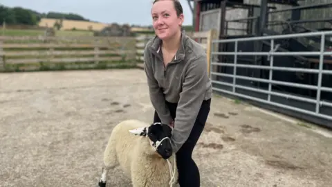 Simon Thake A young girl in a grey fleece holds a sheep carefully with some rope on a farm