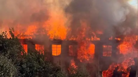 Ginny Adams A burning building with white and grey smoke in the air. Large flames can be seen coming from the building.