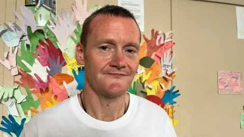 Person wearing a white Nike Air Max T-shirt standing indoors in front of a colourful display of paper handprints with handwritten messages attached to a wall. Several drawings and notices are visible in the background, creating a community or classroom setting.