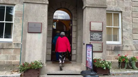 Two people wearing blue and red coats and flat caps walk into Island Hall in Alderney at a previous election. The hall has large grey bricks with four plaques on the exterior wall of the entrance.