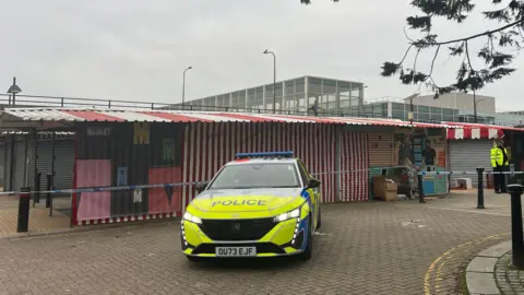 Tony Fisher/BBC Market stalls with a red and white striped canopy can be seen within a blue and white taped police cordon. A police car sits in front of the stalls and a police officer is stood by the side of the cordon. 