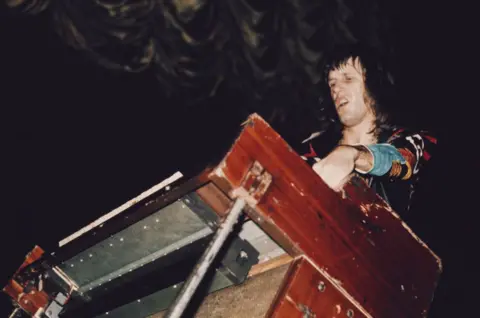 Getty Images Keith Emerson of Emerson, Lake and Palmer plays a battered organ/keyboard, in a flamboyant colourful outfit. He is shot from below, making it look as though he is towering over the instrument.