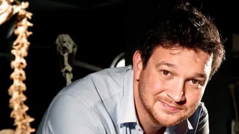 Ben Garrod smiling while looking into the camera. He is wearing a light blue shirt and leaning forward. Next to him appears to be a spine bone. 
