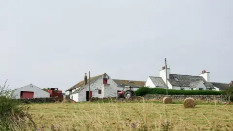 A view of a farm from a distance with white buildings, hay bales in a field and grey skies above
