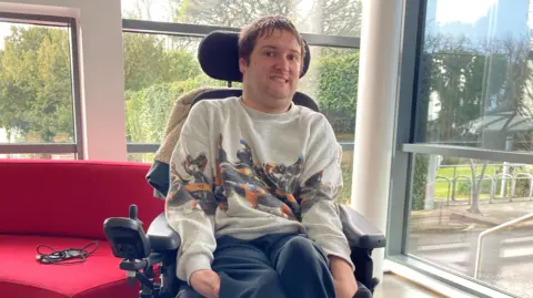 Alex Christmas, who has cerebral palsy, sits in his wheelchair next to a red sofa and several windows. He is wearing a grey jumper with an orange Formula 1 car on it. A mobile phone and a lead are on the sofa.