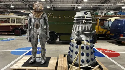 A cyborg and a Dalek model standing in a large warehouse with vintage buses and cars behind