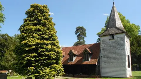 Nigel Cox/Geograph A side-on view of St Andrew's Church, Greensted. On the right is a white-painted wooden tower and its body runs to the left, which is clad in a mix of wood, bricks and flint under a pitched roof. In front of it is grass and an evergreen tree.