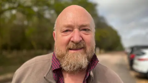 John is wearing a brown jacket with a red and black chequered shirt, and has a ginger-grey beard. 