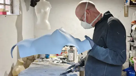 Whitaker Malem Keir Malem in a workshop, holding a white breastplate. He is wearing a white painting mask and a jumper flecked with white paint.
