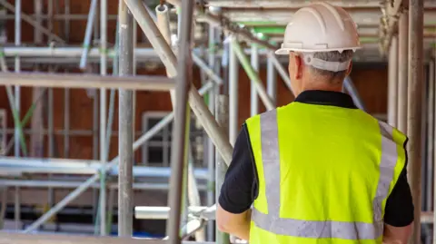 A construction worker, with his back to the camera, is standing in an area of scaffolding. He is wearing a white hard hat and a high visibility vest over a black polo shirt.