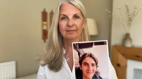 BBC's Anne-Jacques poses for the camera at home wearing a white blouse, a pear-shaped silver necklace and silver earrings. Her long blonde hair is parted on the right. In her right hand, she holds a photo of her late son, Alex Harpum. In the photo, Alex is smiling for the camera with her mouth closed. He has center-parted brown hair, brown eyebrows, and brown eyes. He is wearing a metallic gray down jacket with a hood.