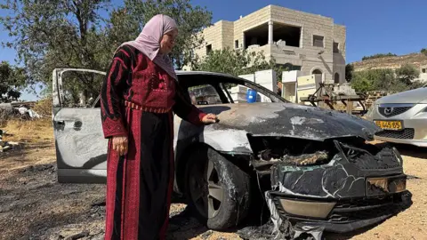 A woman looks at a burned-out car following a settler attack on the Palestinian village of Kafr Malik, near Ramallah, in the occupied West Bank (26 June 2025)