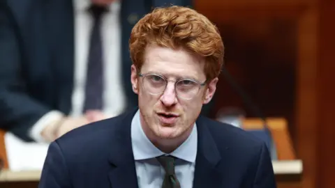 PA Media Matthew O'Toole speaking in the Assembly Chamber during a parliamentary session at Parliament Buildings at Stormont. He has ginger hair and is wearing glasses, a navy suit, patterned green tie and light blue shirt. 