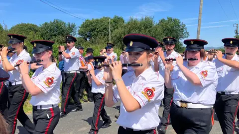 BBC A flute band marching in a parade in County Donegal on a sunny day. They are wearing peaked black caps, white shirts and dark trousers with a red stripe.