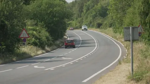 The A36 in Wiltshire. There is a bend in the road and white markings indicating where motorists can overtake and merge. There are other vehicles.