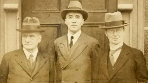 A black and white archive photo of Joseph Fisher, Richard Feetham and Eoin MacNeill during a meeting of the Boundary Commission in Armagh in 1924.  They are all wearing suits. ties and hats and are standing in front of a door.