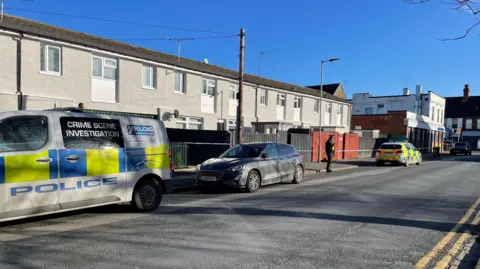 BBC A crime scene investigation van is parked to the left of the photo and police tape is stretched across the front of a row of terraced houses.  A policeman stands in front of the tape.