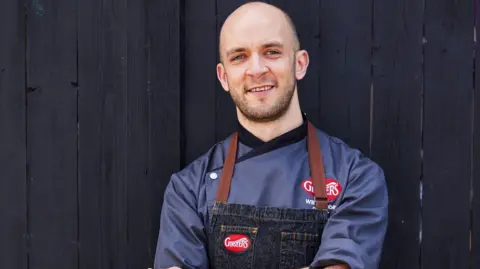 Ginsters A picture of Will wearing a blue chef shirt. He is smiling at the camera - stood in front of a black wall.