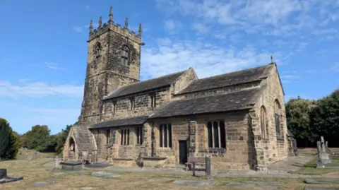 Wakefield Council A large stone church with a square tower and arched windows. Gravestones and flat memorial stones sit on the grass around it, with a few benches nearby.