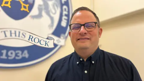 Emma Baugh/BBC A head-and-shoulders picture of Jamie Jones, a middle-aged man with glasses, dark-brown hair and a dark blue shirt. He is standing in a room with cream-coloured walls and, on a wall behind him, part of Peterborough United's crest can be seen.