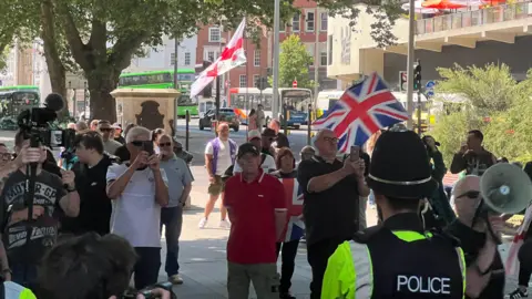 A sparse group of protestors, some of which are holding the England flag and the Union Jack, are shown standing behind a line of police. One person can be seen speaking into a megaphone.