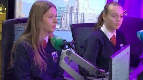 BBC Two girls in school uniforms are in a radio studio in front of microphones. 