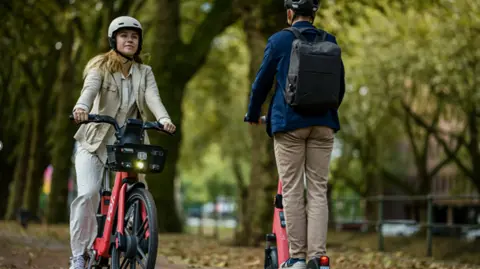 Generic image of a female on an e-bike and male on an e-scooter going passed each other in a park.