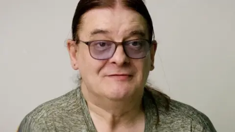 Open House Pictures / UoS Howard, a middle-aged man with long, tied-back hair and tinted glasses. He's stood against a white background, being interviewed for the film.