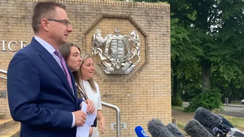 A man with short hair and glasses, wearing a blue suit is in the foreground, standing next to a young woman with long dark hair and a woman dressed in white with long blonde hair. Both women are upset. There are microphones in front of the trio and a court motif on a wall behind them.
