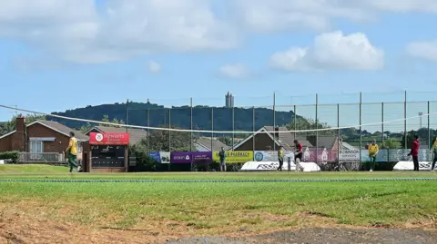 Pacemaker A cricket game being played. Nine layers are on the pitch, some in green and yellow and some in red.