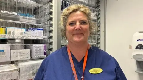 Rachael Conway, dressed in medical scrubs, wearing an orange lanyard and a yellow badge, standing in from of shelves of medication