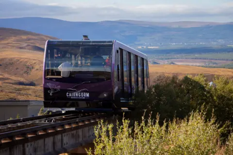 A mountain railway carriage travelling along a steep track. There are rolling hills and trees in the background. The carriage is purple and carrying passengers.