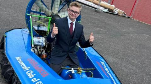 A teenage boy wearing a suit and glasses sitting in the drivers seat of a hovercraft giving two thumbs up